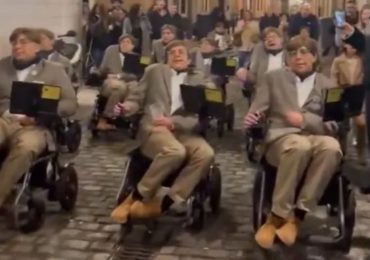 Moment carnival performers dressed as Stephen Hawking parade down street in wheelchairs while singing in robotic voices